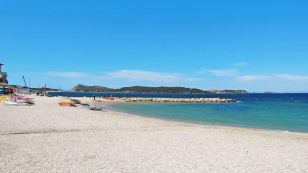 En strand med en vågbrytare av sten, klart blått vatten och en sandstrand.
