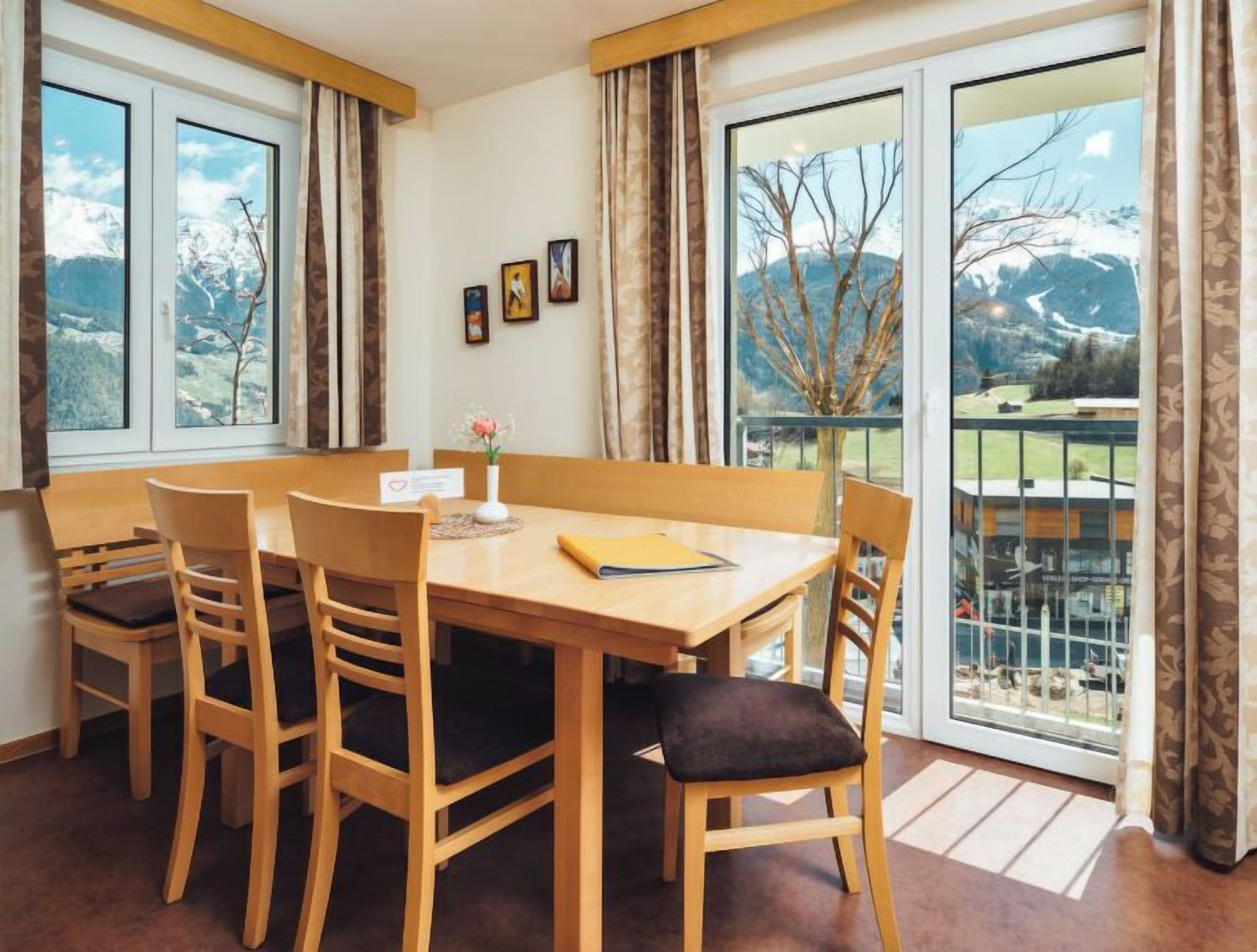 A dining area with a wooden table and chairs, a vase with flowers, and a view of a mountainous landscape through large windows.