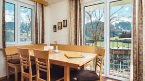 Un coin repas avec une table et des chaises en bois, un vase rempli de fleurs et une vue sur un paysage montagneux, visible à travers de grandes fenêtres.