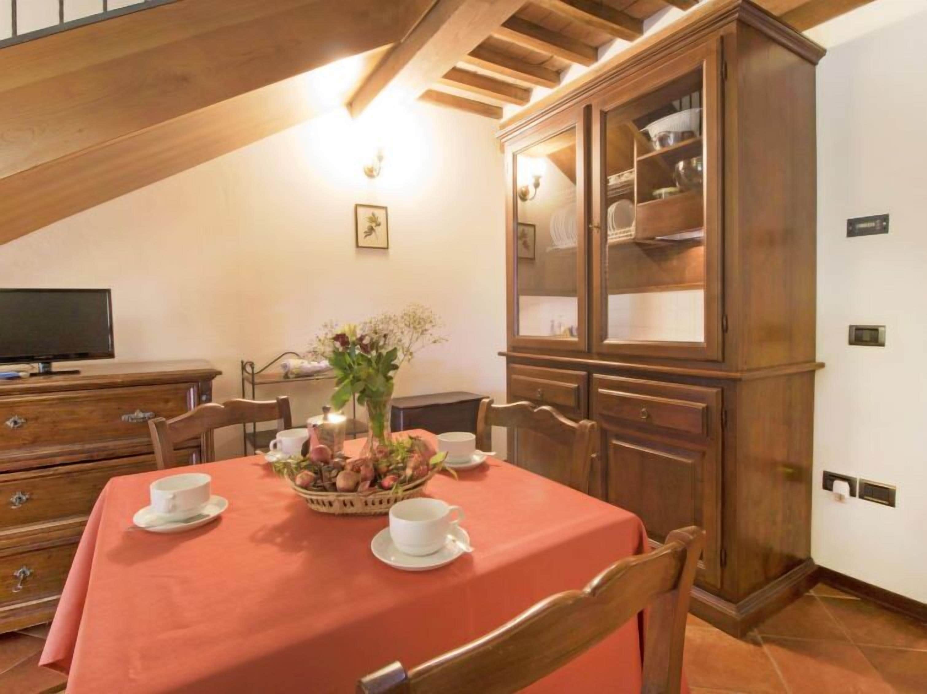A dining area with a table set for two, a cabinet with glass doors, and a television on a dresser.