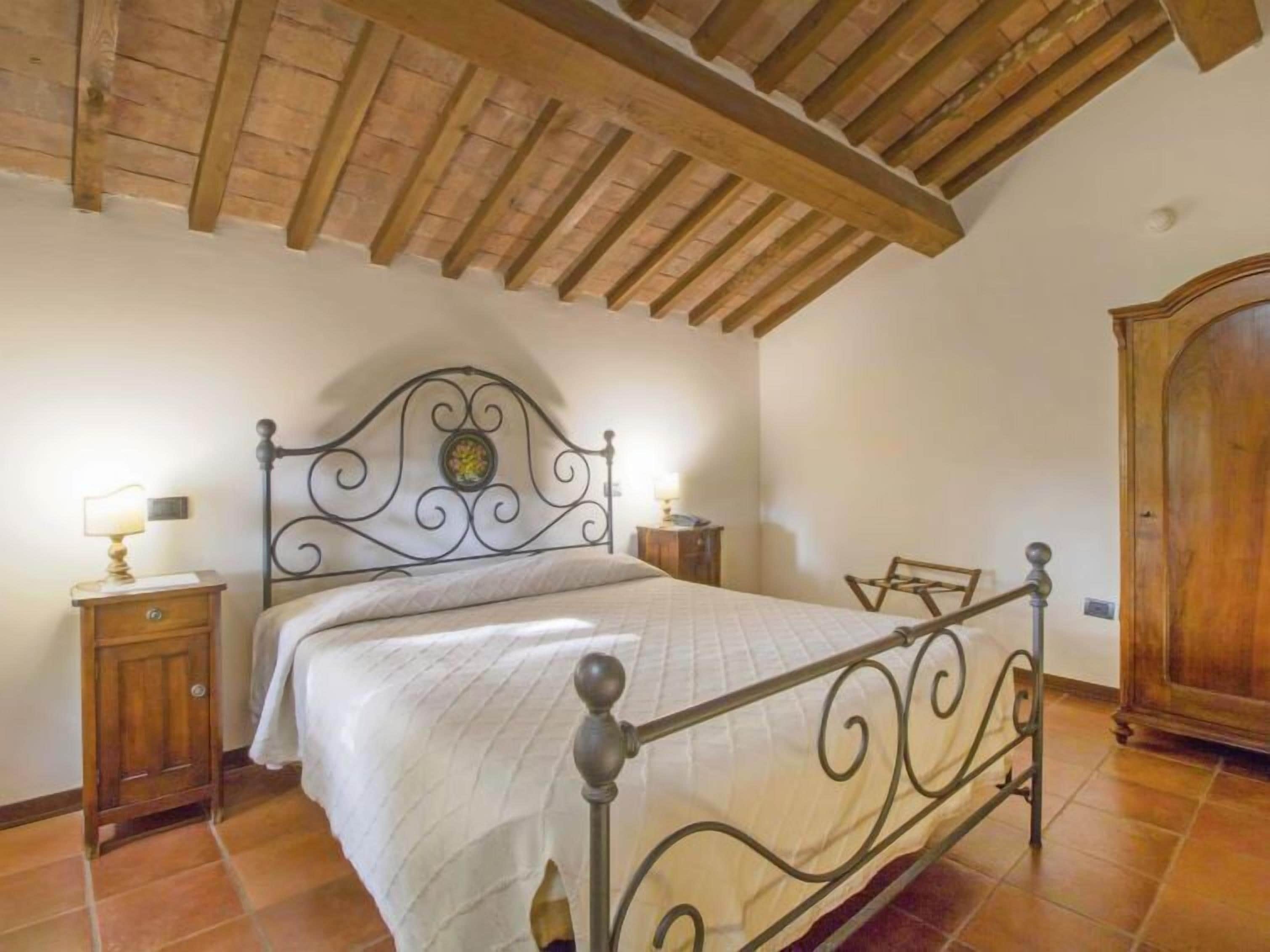 Un dormitorio con estructura de cama de metal, vigas de techo de madera, un armario de madera y una mesita de noche con lámpara.