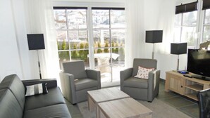 Ein modernes Wohnzimmer mit einem grauen Sofa, zwei Sesseln, einem Couchtisch, einem Fernseher und einem großen Fenster mit Blick auf Gebäude und Bäume.