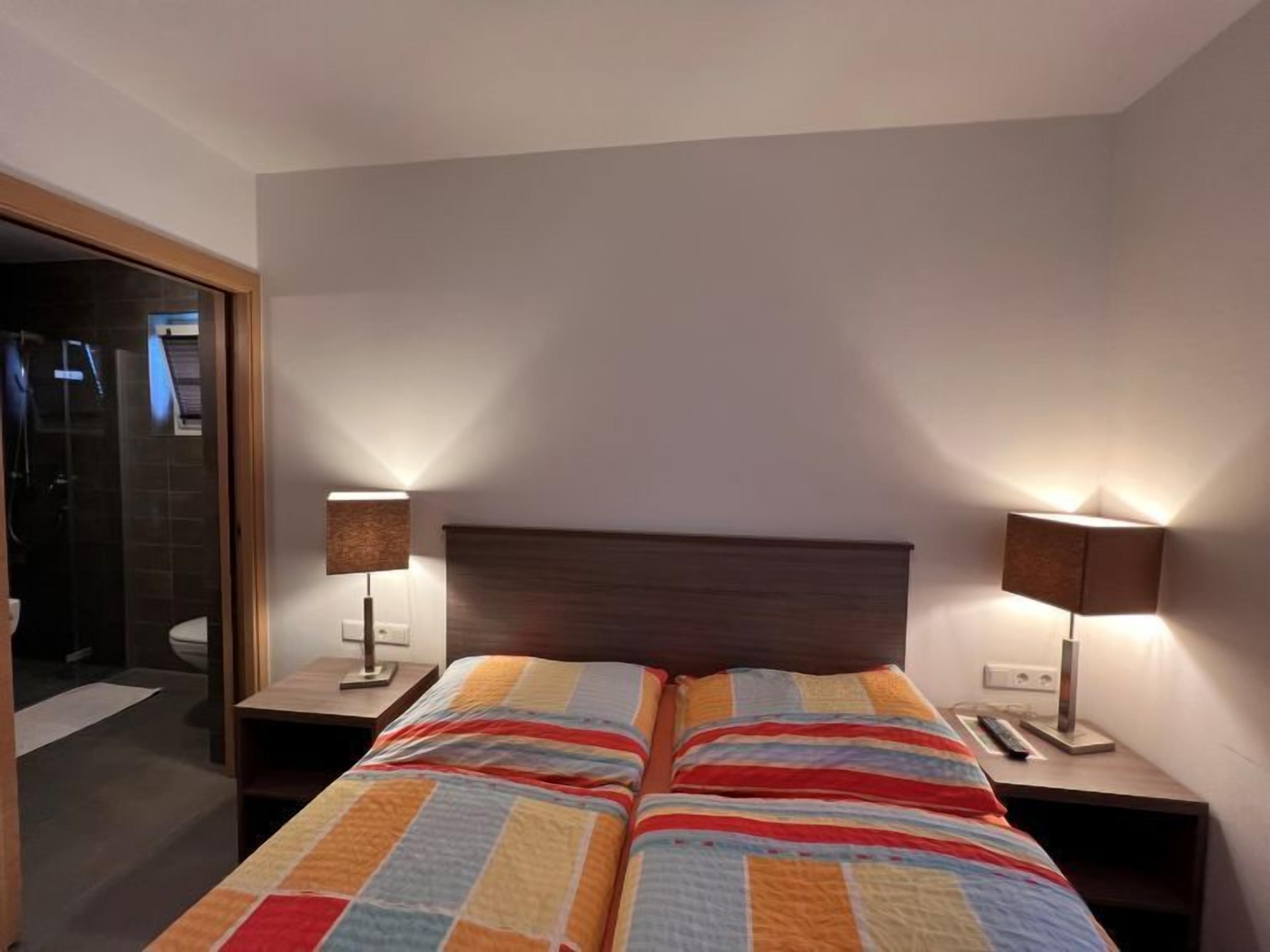 A double bed with a colorful quilt, two bedside tables with lamps, a wooden headboard, and a view of a bathroom with a shower and toilet.