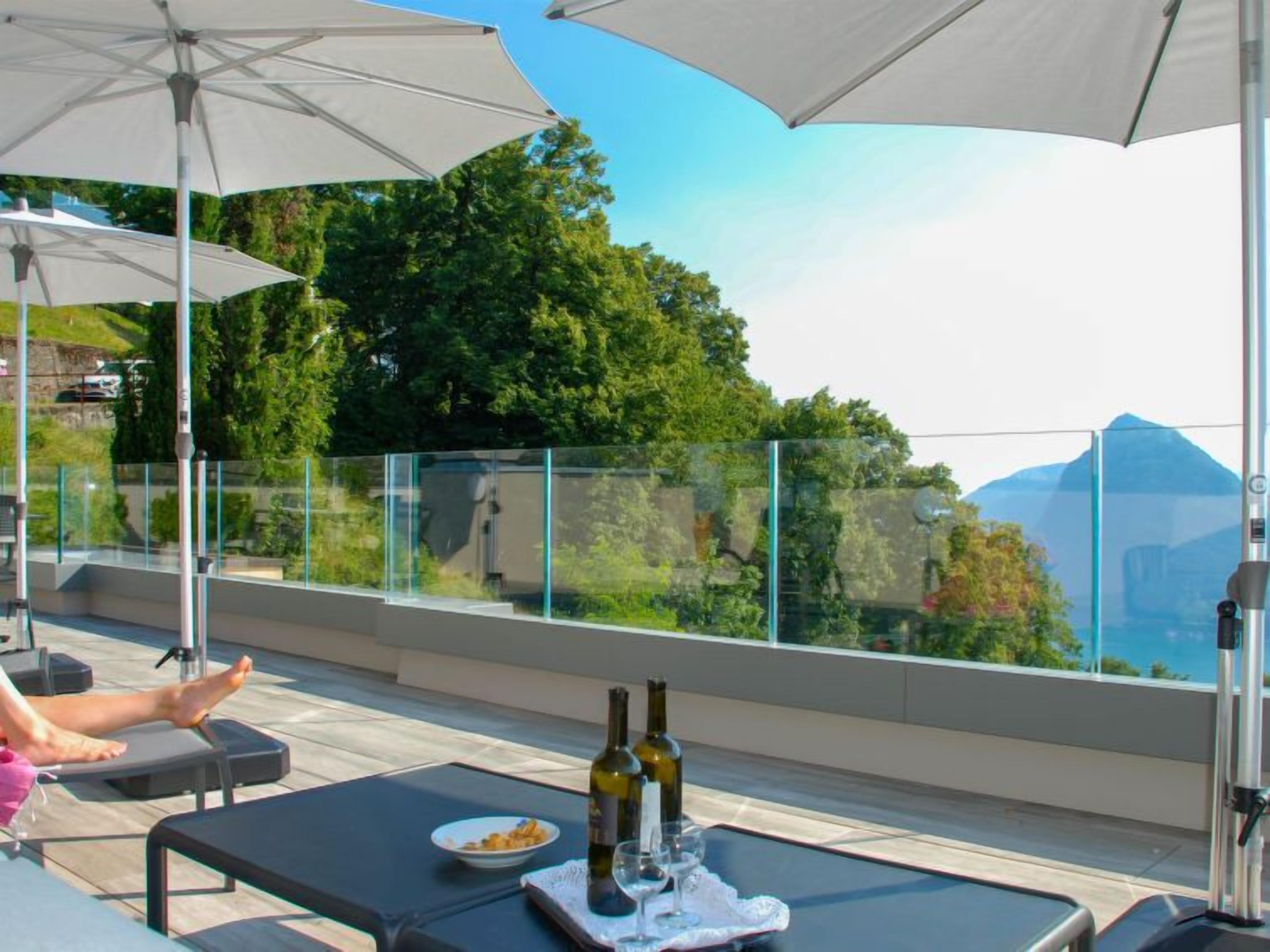 A terrace with outdoor seating, a table with a bottle and glasses, and a view of greenery and mountains.