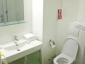 A bathroom with a white sink, mirror, and toilet. There is a glass shelf above the sink and a stack of white towels on the wall. - Residenza Monte Bré - Apt.105 Haus1 by Interhome (Aldesago)