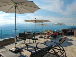 A rooftop terrace with outdoor seating, umbrellas, and a panoramic mountain view. - Residenza Monte Bré - Apt.105 Haus1 by Interhome (Aldesago)