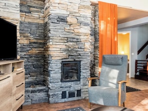 A stone fireplace with a television on a wooden cabinet, a grey armchair, and a wooden staircase.