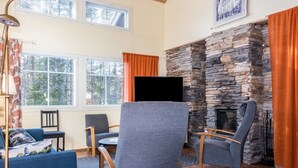 A cozy living room with a stone fireplace, comfortable seating, and large windows offering a view of the outdoors.