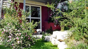 A modern house with a red exterior, a balcony with a metal railing, and a garden with pink flowers and greenery.