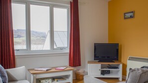 A living room with a sofa, a TV on a stand, a window with curtains, and a view of the outdoors. - Alan's House by Interhome (North Skye)