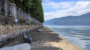 A concrete waterfront promenade with a stone wall and metal railing, overlooking a calm lake and distant mountains.