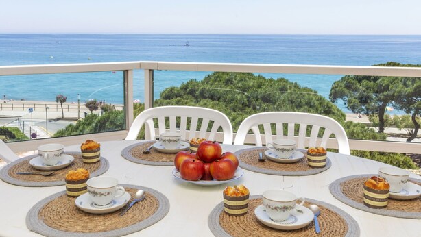 A table set for breakfast with a view of the sea, white chairs, and a balcony railing.