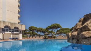 A swimming pool with lounge chairs and umbrellas, surrounded by trees and a building with balconies.