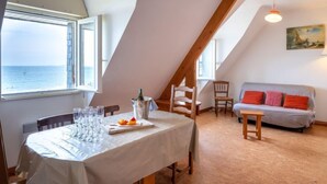 A dining area with a table set for four, a view of the sea through a window, and a loft-style room with a sofa and a painting. - Vacation home Trezen by Interhome (Carnac)