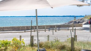 An outdoor dining area with a view of the sea, tables with breakfast items, and a person walking along the rocky shore. - Vacation home Trezen by Interhome (Carnac)