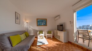 Un salon avec un canapé gris, un fauteuil blanc, une table basse et un téléviseur posé sur un meuble blanc.