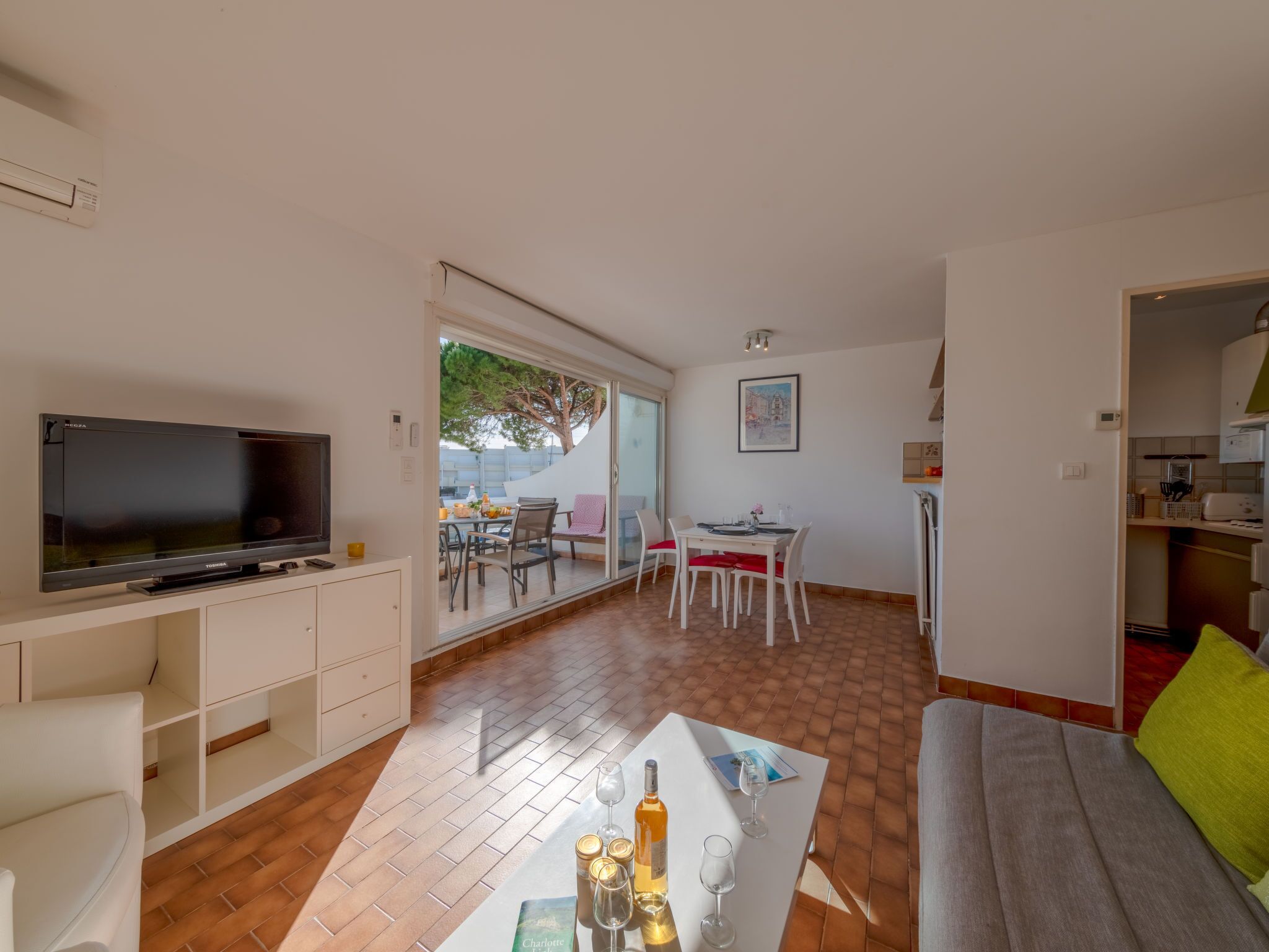 A modern living room with a flat-screen TV, a dining area with a table and chairs, and a kitchen area with cabinets and appliances.