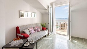 Una sala de estar moderna con un sofá gris, cojines rojos, una mesita con mantel de encaje y vista al mar desde un balcón abierto.