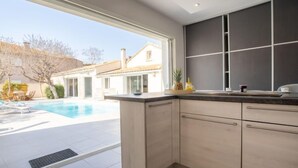 Eine moderne Küche mit einem großen Fenster, einer integrierten Kochinsel und Blick auf einen Swimmingpool sowie benachbarte Häuser.
