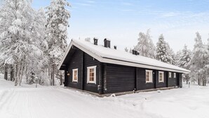 Lumipeitteinen, jyrkkäkattoinen puumökki, jonka ympärillä on lumikuormaisia puita.