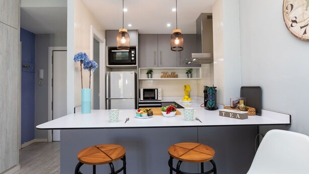 A modern kitchen with a central island, two bar stools, a microwave, a refrigerator, and a clock on the wall.