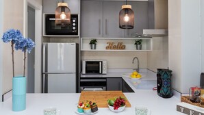A modern kitchen with a refrigerator, microwave, and oven. There are two hanging pendant lights, a sink with a bowl of lemons, and a coffee machine.