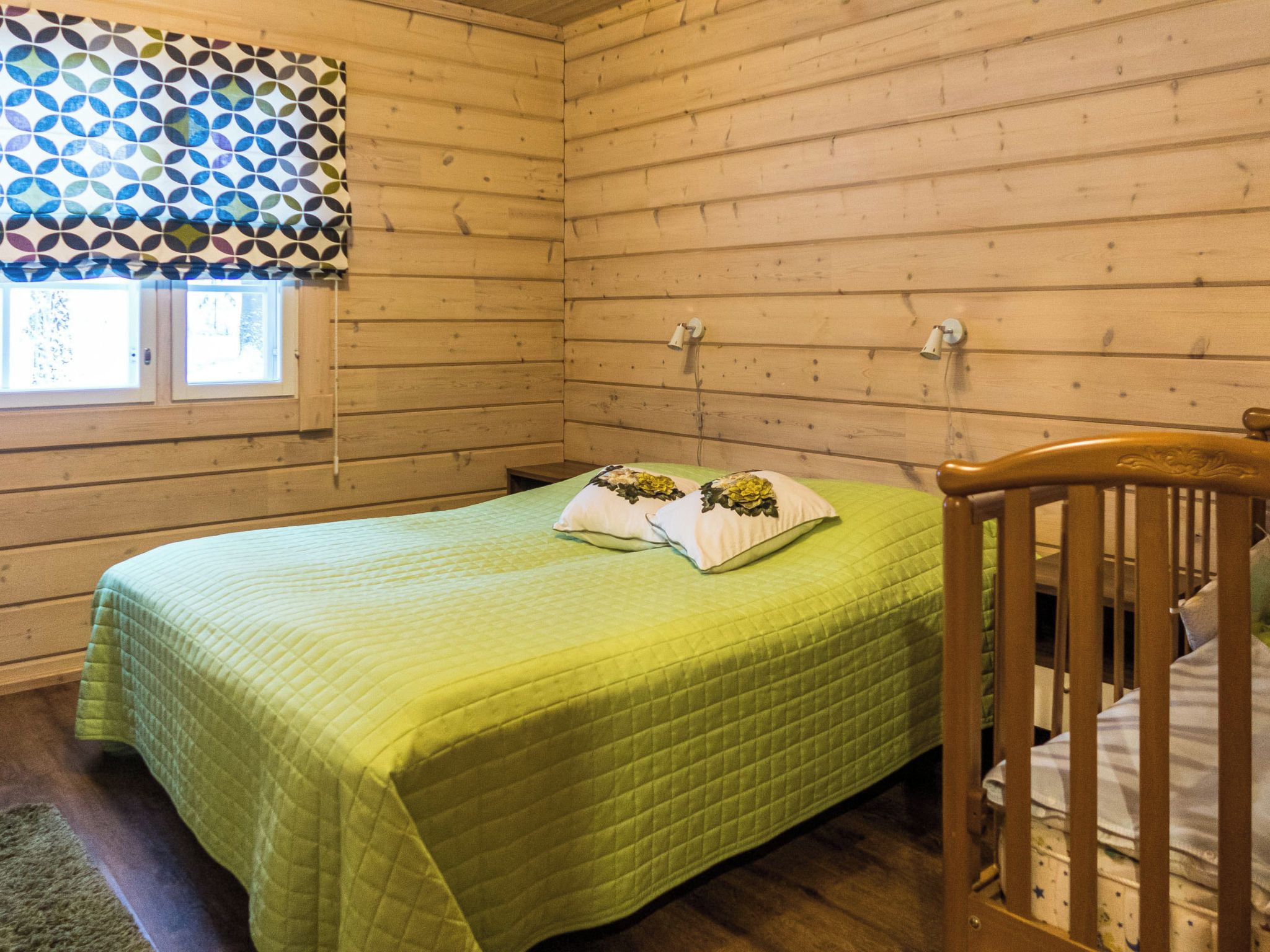 Una habitación con una cama, una cuna, un ventanal con cortinas estampadas y una pared de madera.