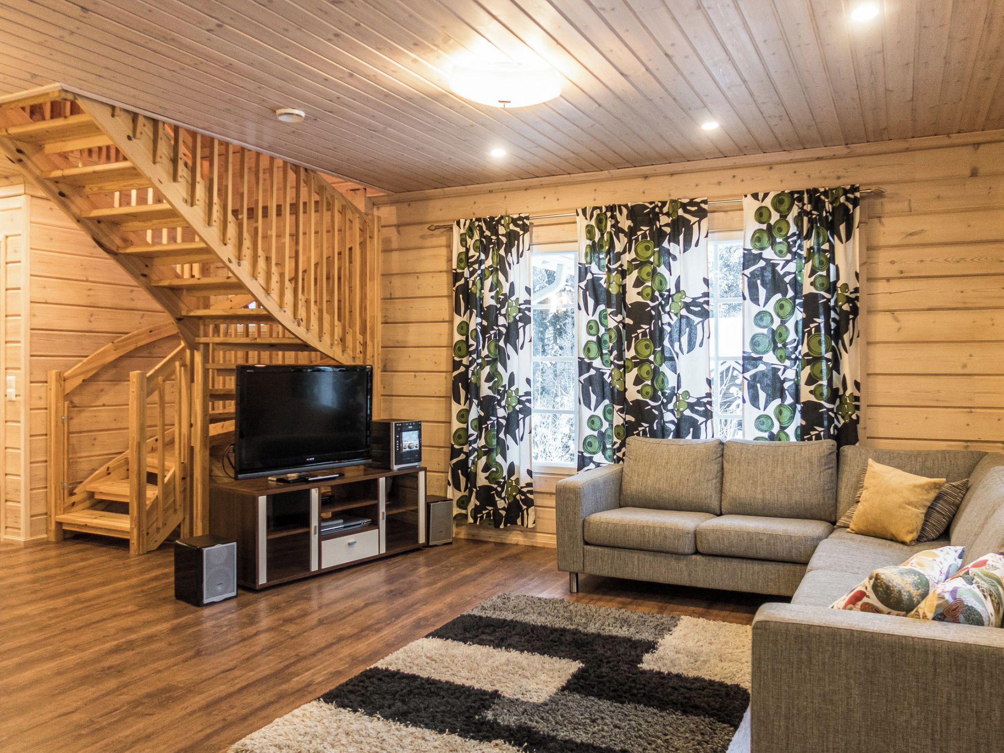 Interior em madeira com escada, televisão em um armário, sofá e cortinas estampadas.