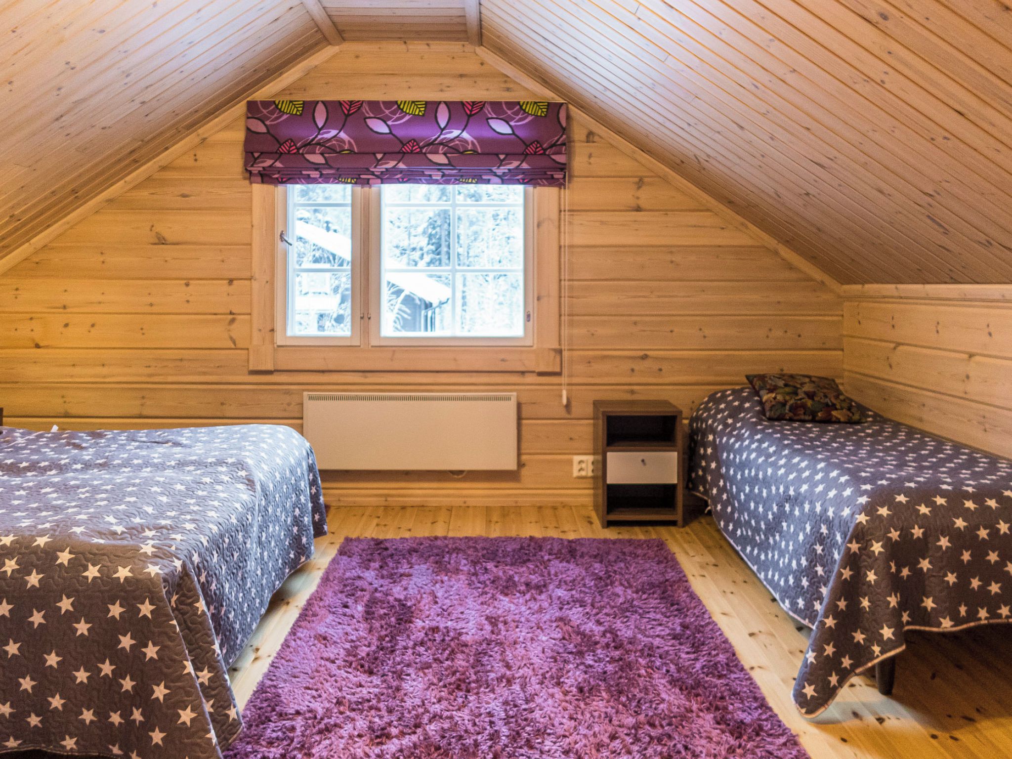 A cozy attic bedroom with two beds, a purple rug, and a window with a leaf-patterned valance.