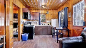 Cocina compacta con armarios de madera, microondas, estufa y refrigerador. Se aprecia una ventana con persianas y una pequeña mesa con sillas.