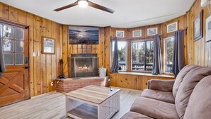 Ein gemütliches Blockhaus-Interieur mit Kamin, Holzwänden, Deckenventilator und einem großen Fenster mit Blick auf eine Terrasse.