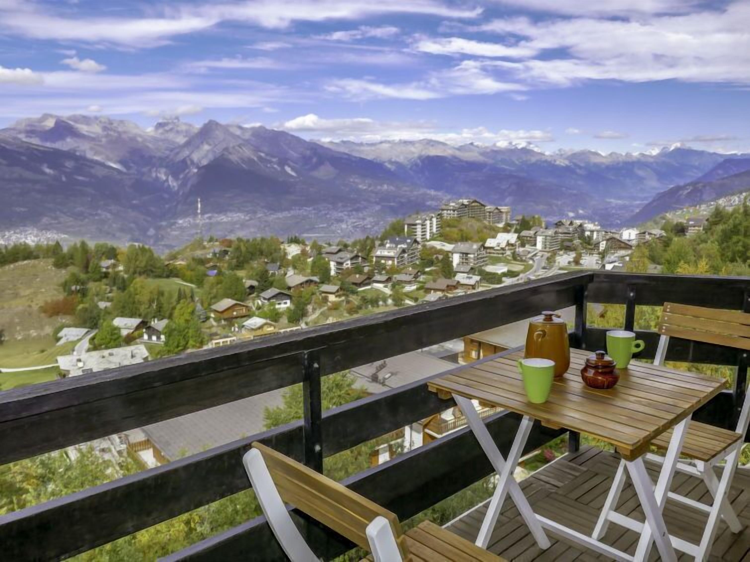 Une terrasse en bois avec une table et des chaises, d’où l’on a une vue sur un village de montagne avec ses maisons et son église.