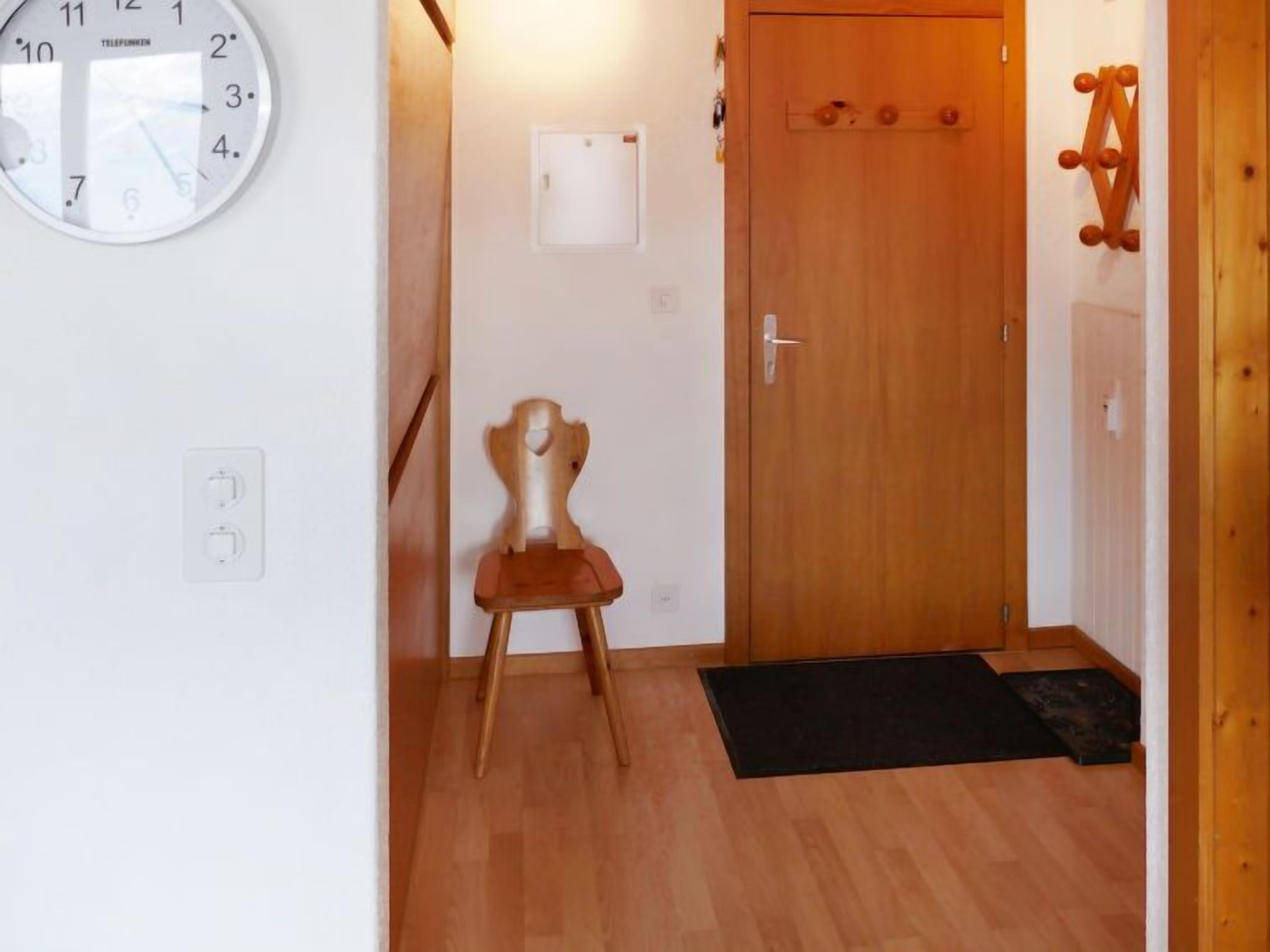 Un pasillo con una puerta de madera, un reloj en la pared y una pequeña silla de madera.