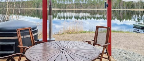 Una terraza de madera con una mesa redonda y sillas, con vista a un lago tranquilo y árboles al fondo.