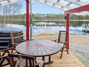 En træterrasse med et rundt bord og stole, med udsigt over en rolig sø og træer i baggrunden.