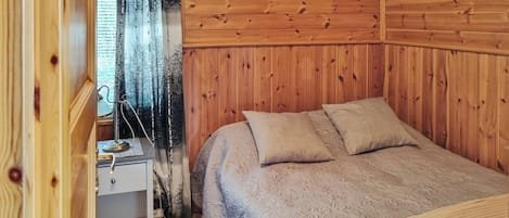 A cozy room with a wooden paneled wall, a bed with a patterned bedspread, a nightstand, and a window with a curtain.