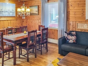 Et rum med træpaneler, et spisebord, stole, en lysekrone, en sofa og et vindue med gardiner.