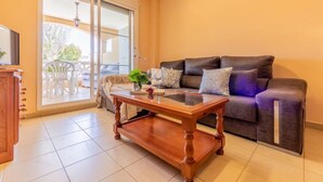 A living room with a dark brown sofa, a wooden coffee table, and a sliding glass door leading to an outdoor area with a white table and chairs. - Res Mediterraneo by Interhome (Deltebre)