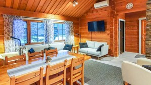 L’intérieur chaleureux d’un chalet avec une table à manger en bois, deux canapés et un téléviseur fixé au mur.