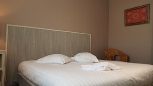 A neatly made bed with white linens and a folded towel, a wooden headboard, and a wicker chair in a room with a plain wall and a framed picture. - furnished apartment with all mod cons in a detached house with a garden (Bagnoles-de-l'Orne)