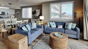 A living room with two sofas, a dining area with a table and chairs, a kitchen with appliances, and a large window with blinds. - Sunrise Views at Emerald Key 201 – Gulf Views! (Orange Beach)
