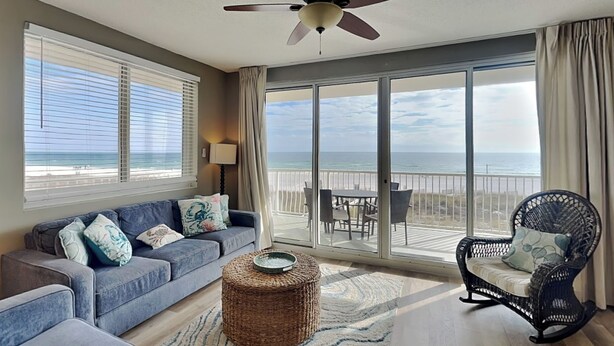 A spacious living room with a large sofa, a wicker ottoman, a wicker chair, and a ceiling fan. A balcony with a view of the beach is visible through large windows. - Sunrise Views at Emerald Key 201 – Gulf Views! (Orange Beach)
