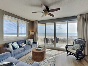 Uma sala de estar espaçosa com um sofá grande, um banquinho de vime, uma poltrona de vime e um ventilador de teto. É visível uma varanda com vista para a praia através de amplas janelas.