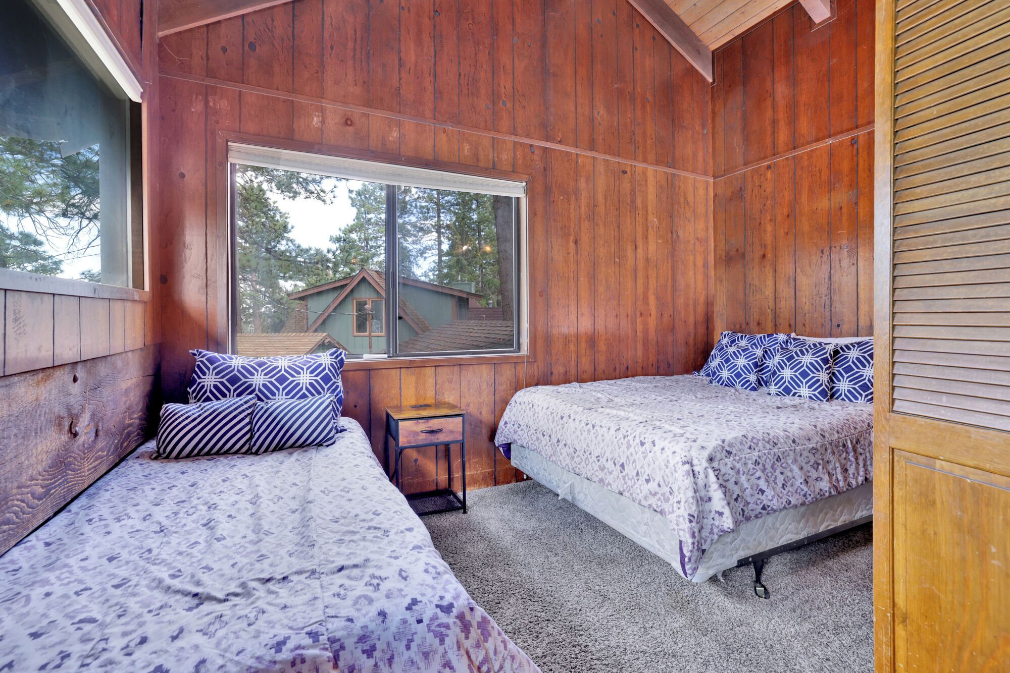 Un dormitorio de cabaña de madera con dos camas, una mesita de noche de madera y un ventanal que ofrece vistas a una casa y árboles.
