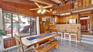 A rustic cabin interior with a wooden dining table, benches, a kitchen area with wooden cabinets, and a large window overlooking a backyard.