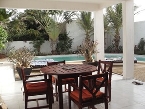 An outdoor patio with a round wooden table and chairs, a pool, and various plants.