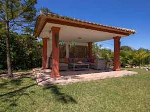 A covered outdoor seating area with a sofa and table, surrounded by greenery and trees.
