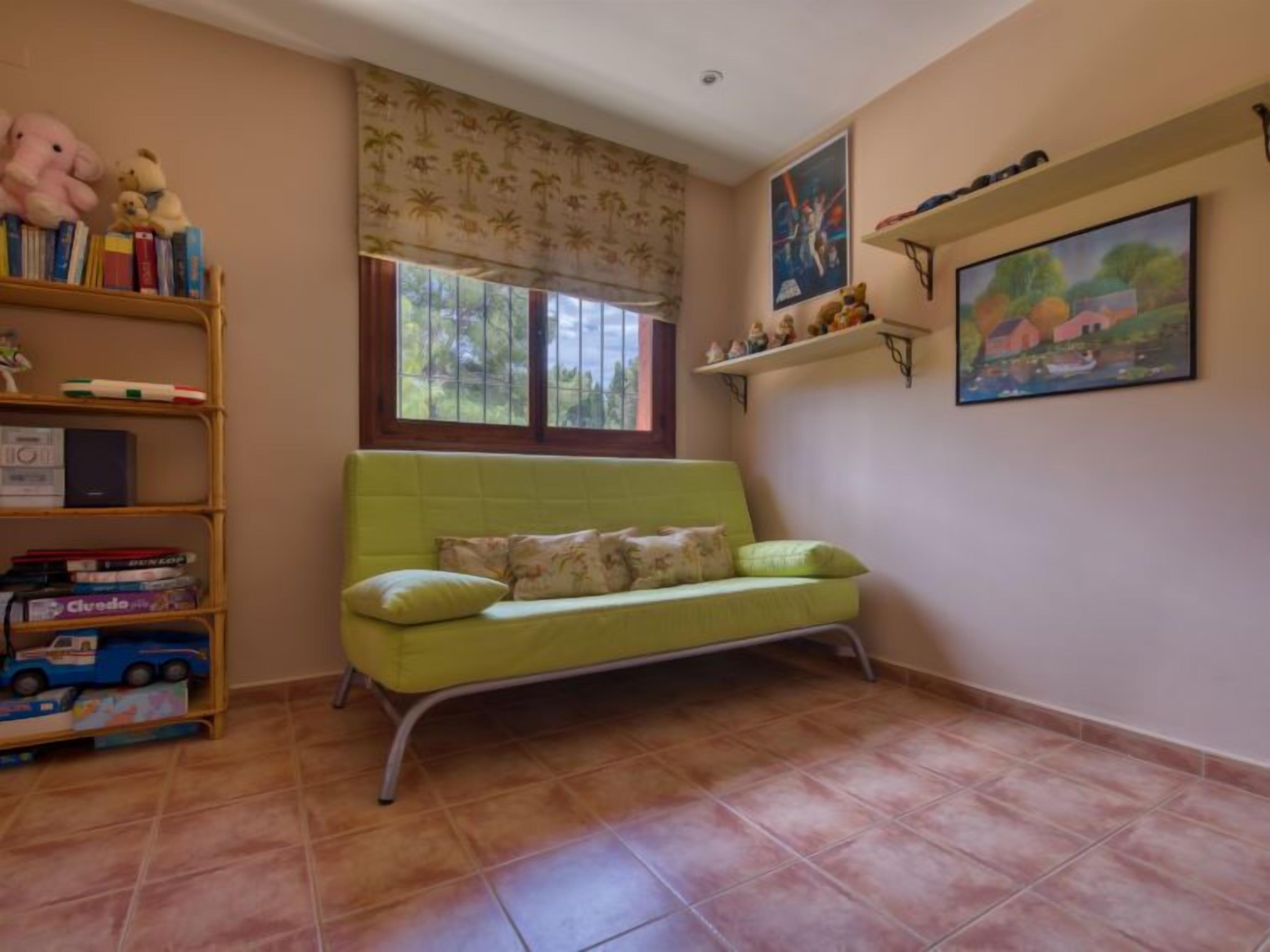 Een kamer met een groene bank, een boekenkast met boeken en speelgoed, een raam met uitzicht op bomen, en twee wandplanken met decoratieve objecten.