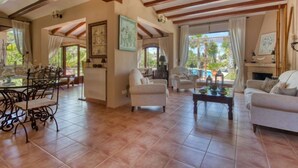 Ampla sala de estar com teto alto, piso em azulejo e vista para piscina. Possui vários espaços de estar, incluindo uma mesa de jantar com cadeiras e um sofá.