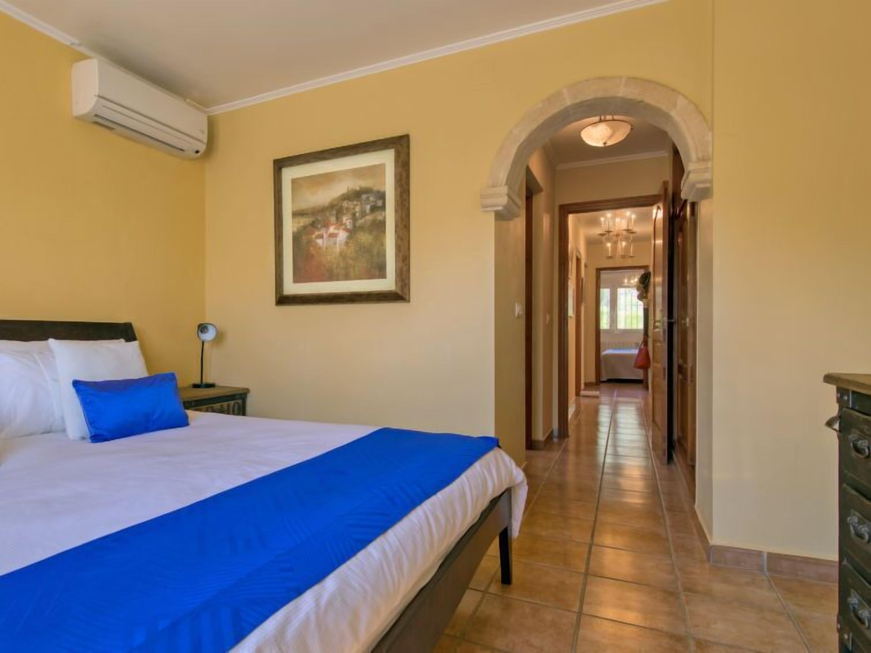 A bedroom with a bed, a nightstand, a lamp, a painting, and a hallway leading to another room.